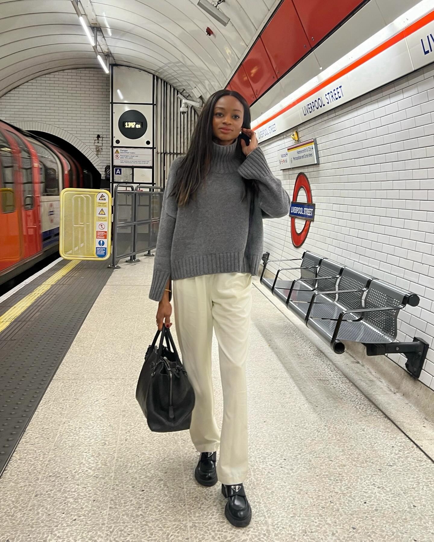 British style influencer Lorna Humphrey poses in the London Underground. wearing a gray turtleneck sweater, ivory relaxed silk pants, a black bag, and black Prada loafers