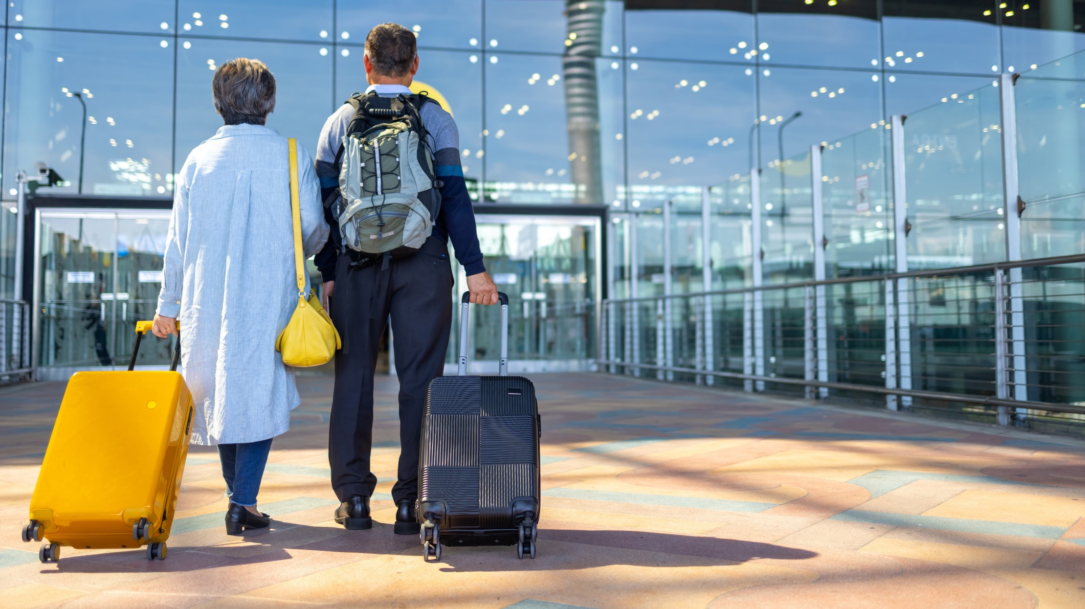 A senior couple wheeling luggage while walking into an airport terminal.