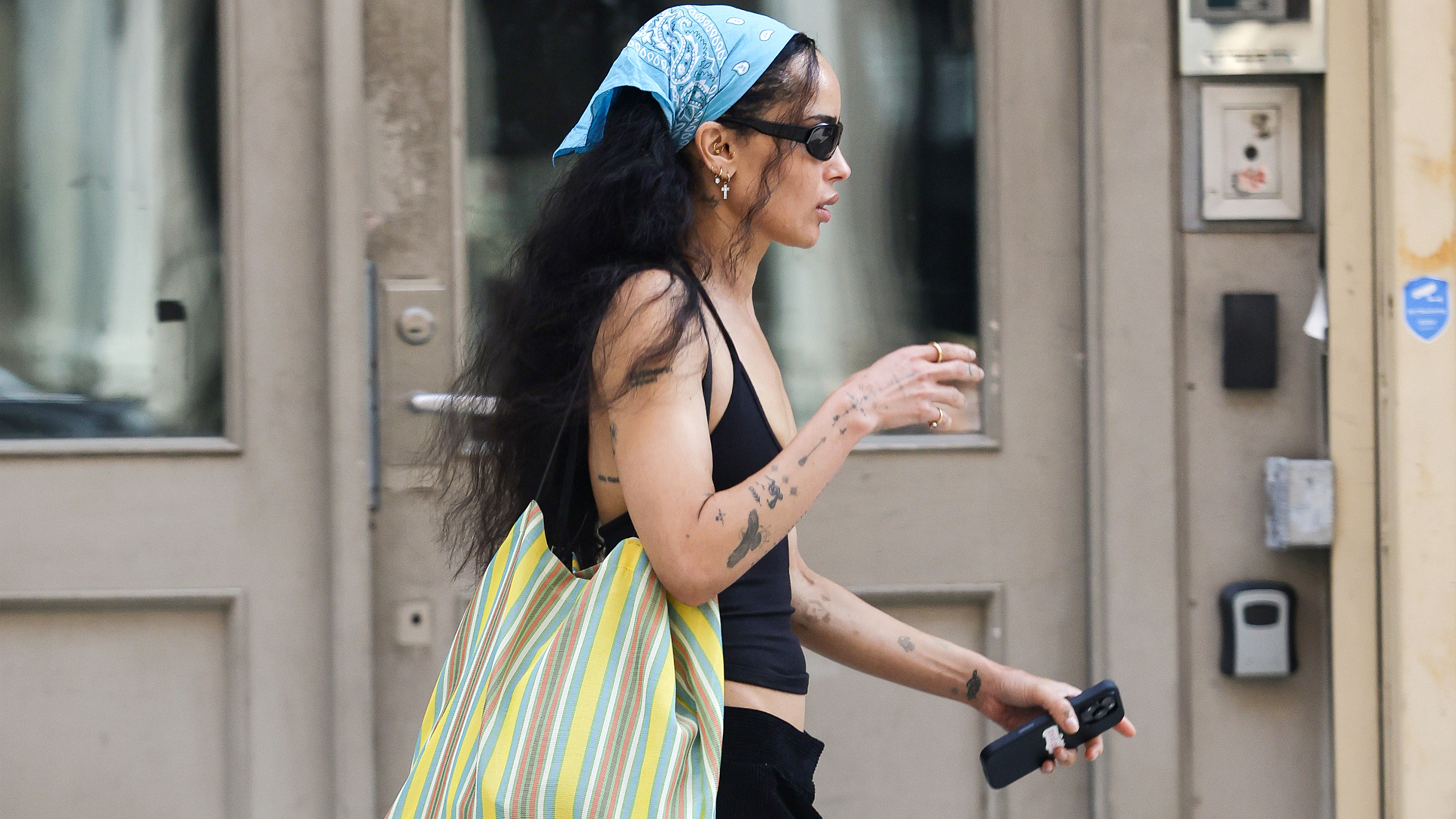 Zoe Kravitz flashes a smile while stepping out in New York City. The American actress carried a large striped tote bag and wore a bandana, black tank top, matching trousers, and sandals.