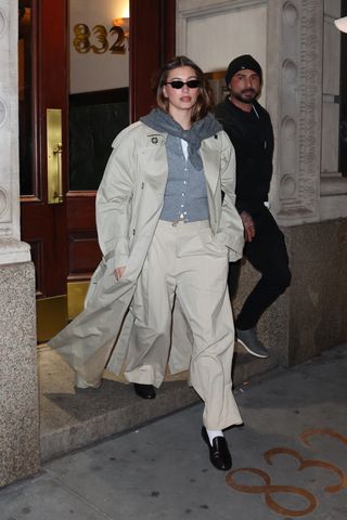 Hailey Bieber wears a trench coat, gray cardigan, gray scarf, beige trousers, socks, and loafers.