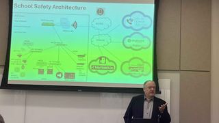 A professor discusses school safety systems in front of a screen.