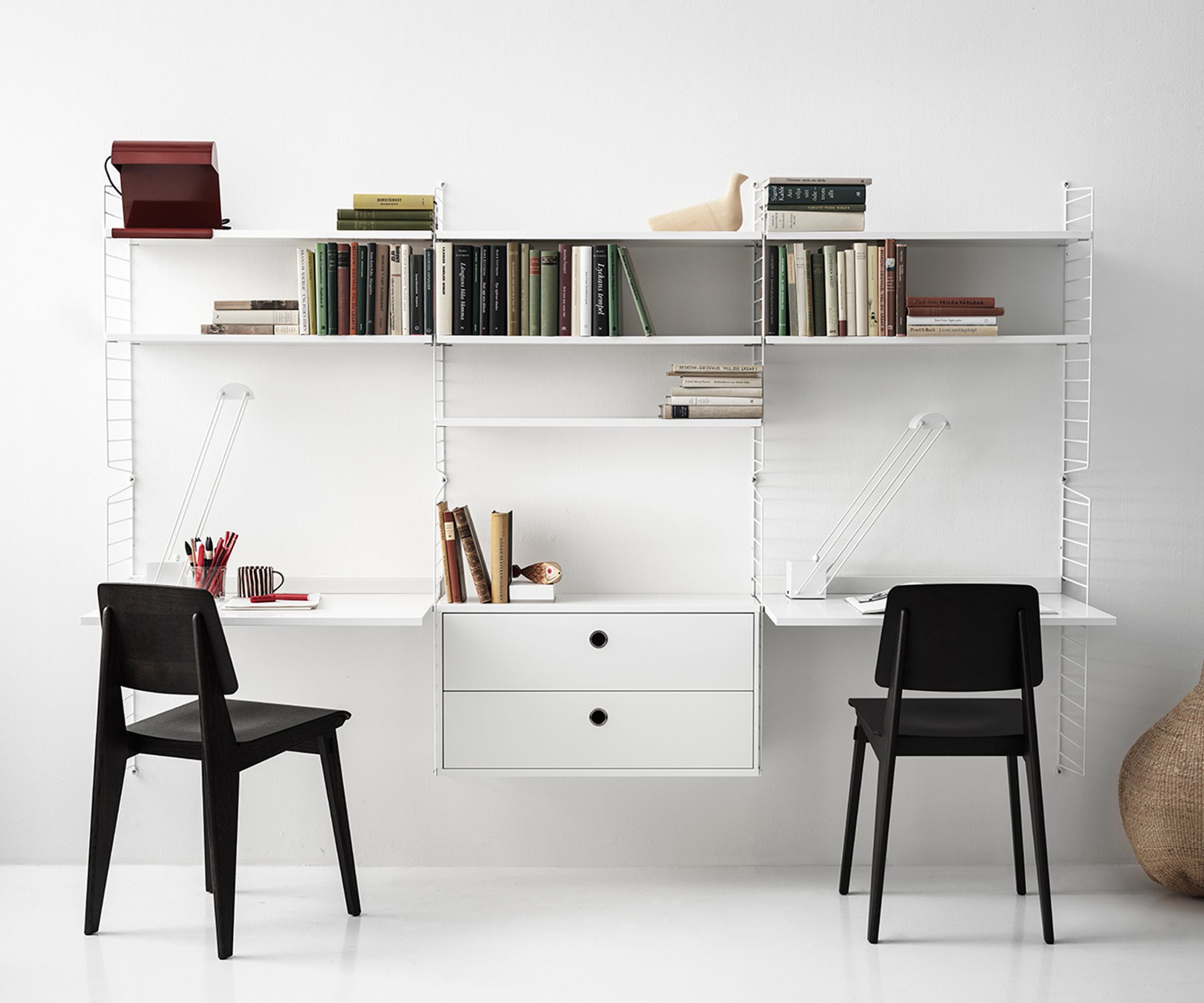 Two minimalist desks with black chairs sit beneath white wall-mounted shelving filled with books and decor, creating a tidy dual workstation.