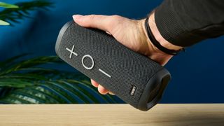 a close up picture of the three volume control buttons of a black oblong-shaped bluetooth speaker with mesh covering and a carry strap photographed on a beige table in front of a navy wall