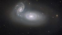 Two wispy white galaxies swirl next to each other in black space, with some faint stars and distant galaxies scattered about.