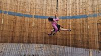 Picture of a female runner wearing a long-sleeve pink top, black shorts and purple and black trainers. She is running along a "Wall of Death" which appears to be made of wood. There is a blue stripe of paint running across the circumference of the wall. She is also holding on to bungee cords and is wearing a harness.