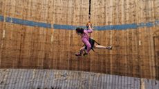 Picture of a female runner wearing a long-sleeve pink top, black shorts and purple and black trainers. She is running along a "Wall of Death" which appears to be made of wood. There is a blue stripe of paint running across the circumference of the wall. She is also holding on to bungee cords and is wearing a harness.