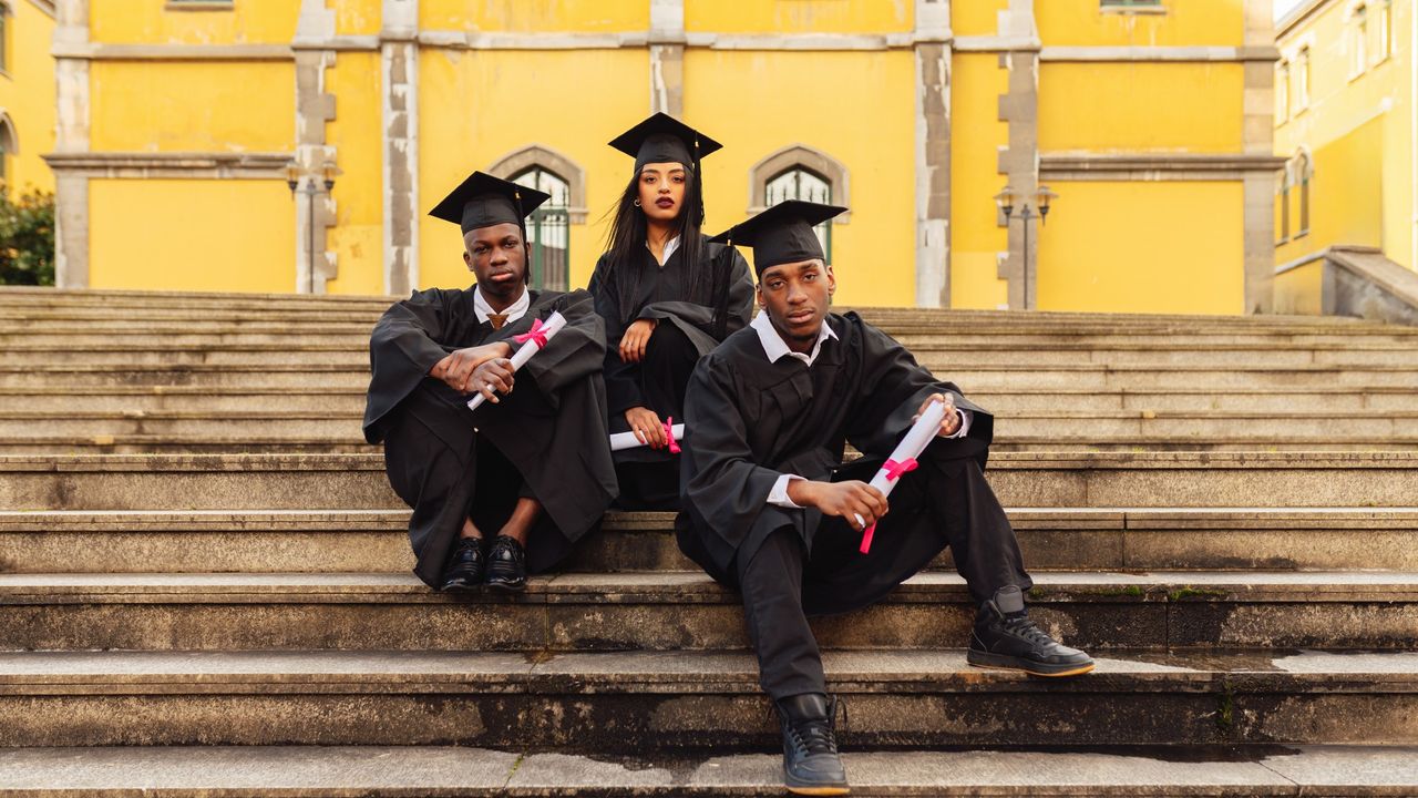 Three young students sitting on university steps, wearing graduation gowns and holding diplomas