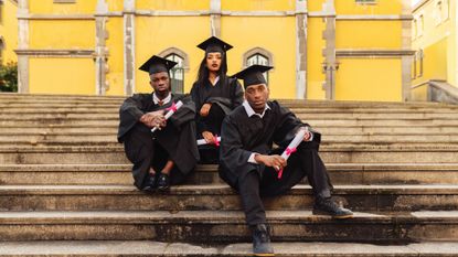Three young students sitting on university steps, wearing graduation gowns and holding diplomas