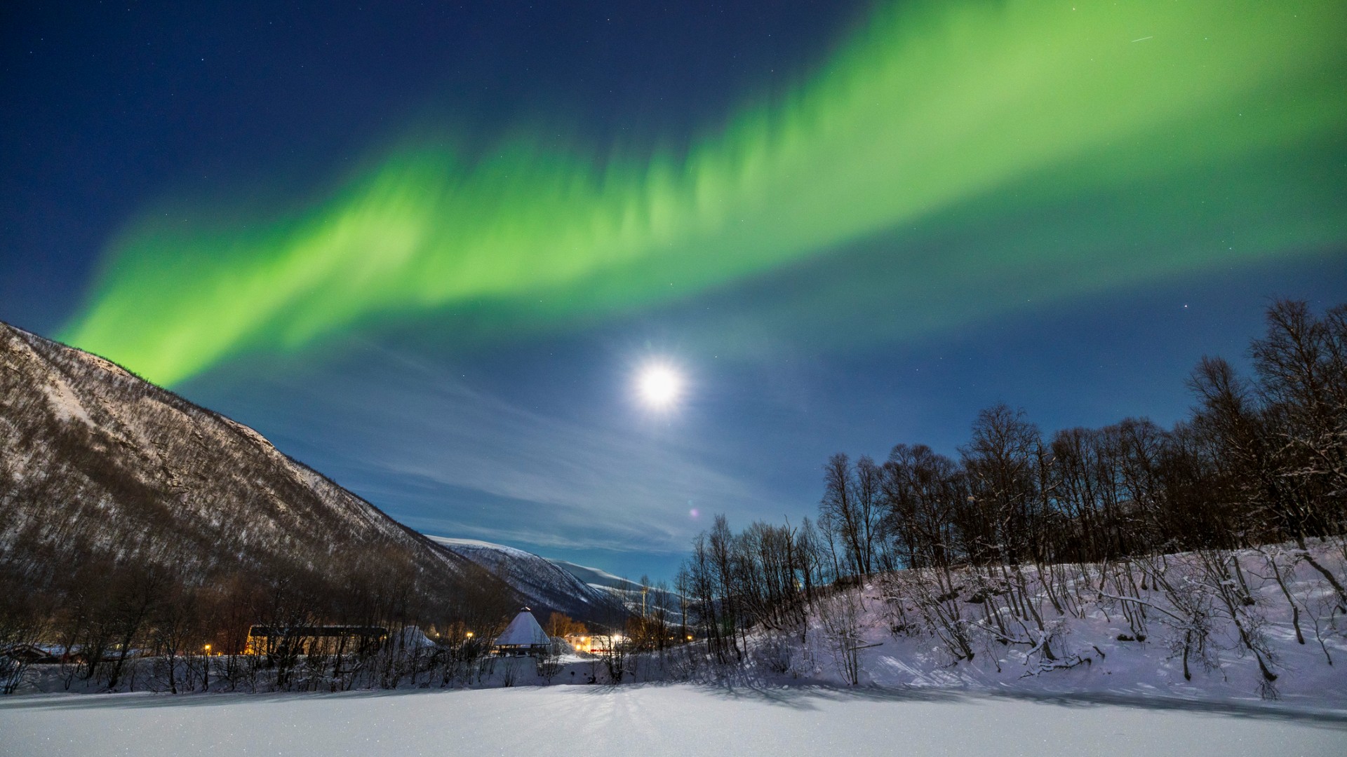 Green northern lights arc over snowy mountains and trees at night, with a bright moon in a clear sky above a small settlement.
