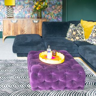 A living room with a navy velvet corner sofa and a purple quilted ottoman instead of a coffee table