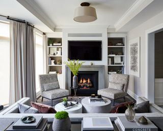 Family room space with gray painted walls and cozy seating area, black flooring with gray rug, two gray upholstered lounge chairs, dark gray sofa facing the fireplace and tv with cushions, built in shelving in alcoves beside the fireplace, gray curtains and beige pendant light, two rounded low gray coffee tables
