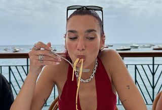 Dua Lipa in Italy eating pasta wearing a red halterneck top with black sunglasses.