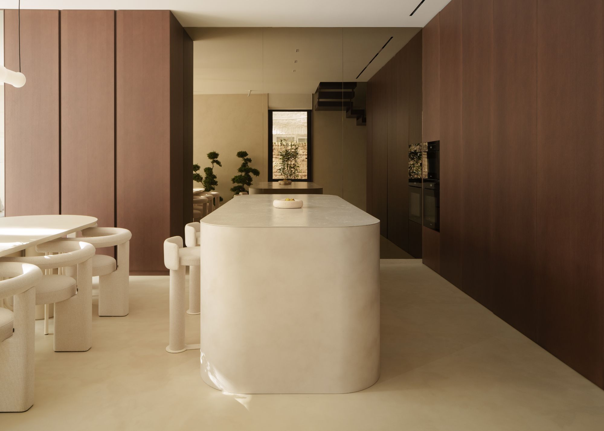 A rounded, cream-colored marble kitchen island with cream bar stools in front of a mirrored wall, with a curved cream dining table just out of view, and surrounded by wood units