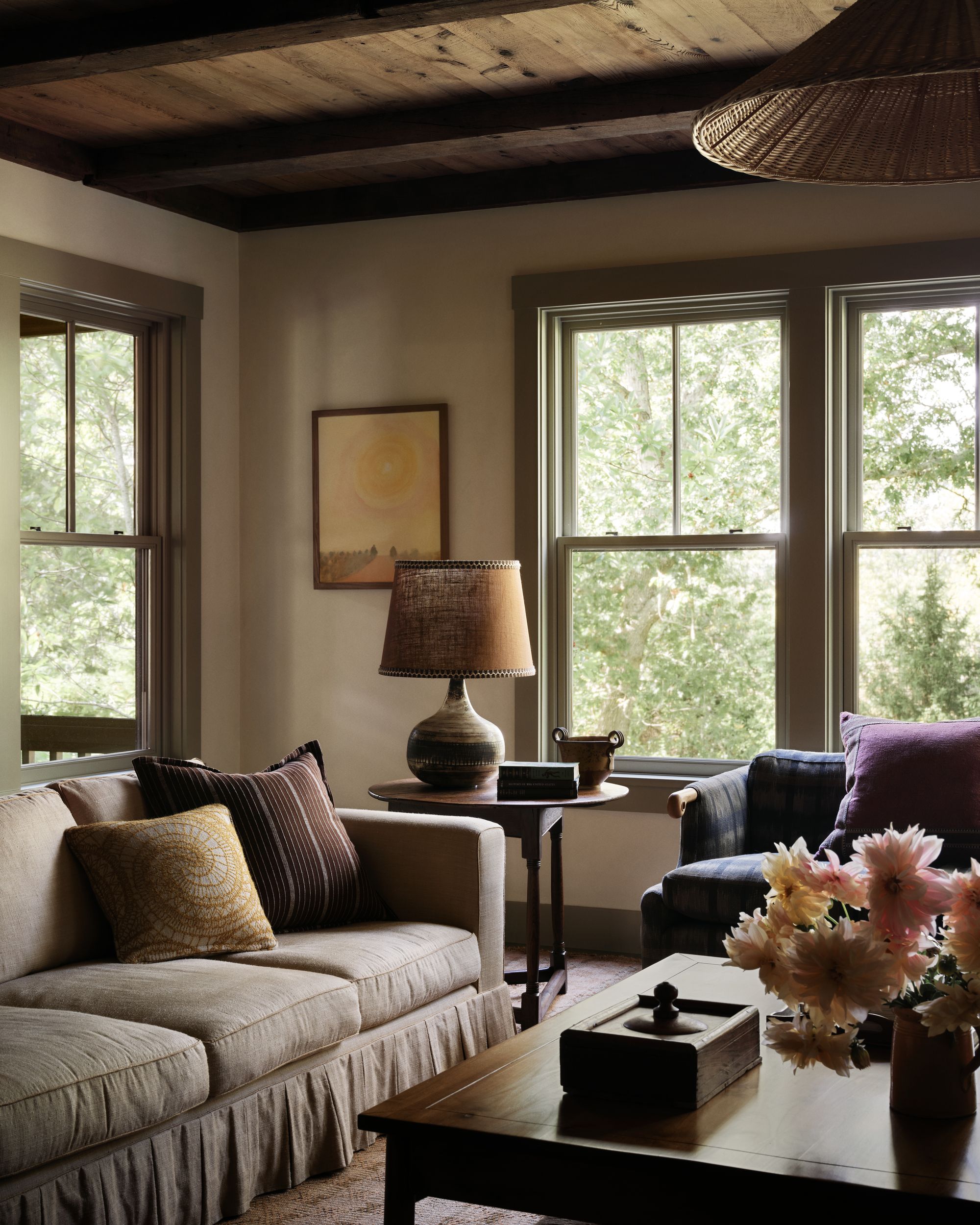 Image of a cozy living room with a dark, neutral color palette. There is a beige skirted sofa, a wooden side table with a large lamp, a blue plaid sofa, and a wooden coffee table.