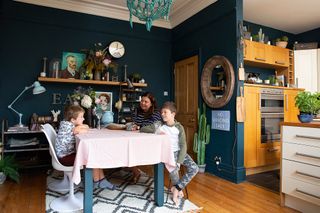 The family sit around the dining table. The room has dark navy walls and there is a turquoise chandelier hanging from the ceiling. The grey table is covered with a light pink tablecloth