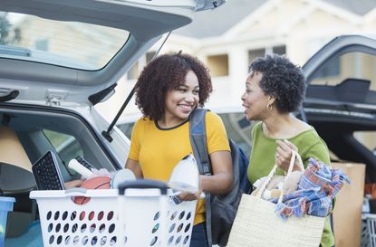 A mature African-American woman helping her daughter relocate, perhaps into an apartment or college dorm.They are in a parking lot unloading their cars. The young woman is carrying a laundry 