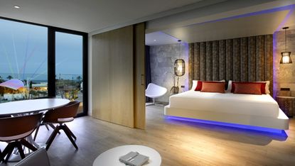 Hard Rock Hotel room, light wood floor, white double bed with blue light underneath, white chair, dining table and chairs, black frame window and balcony door, view of surrounding area, stone coloured and patterned walls, wall lights, round white coffee table