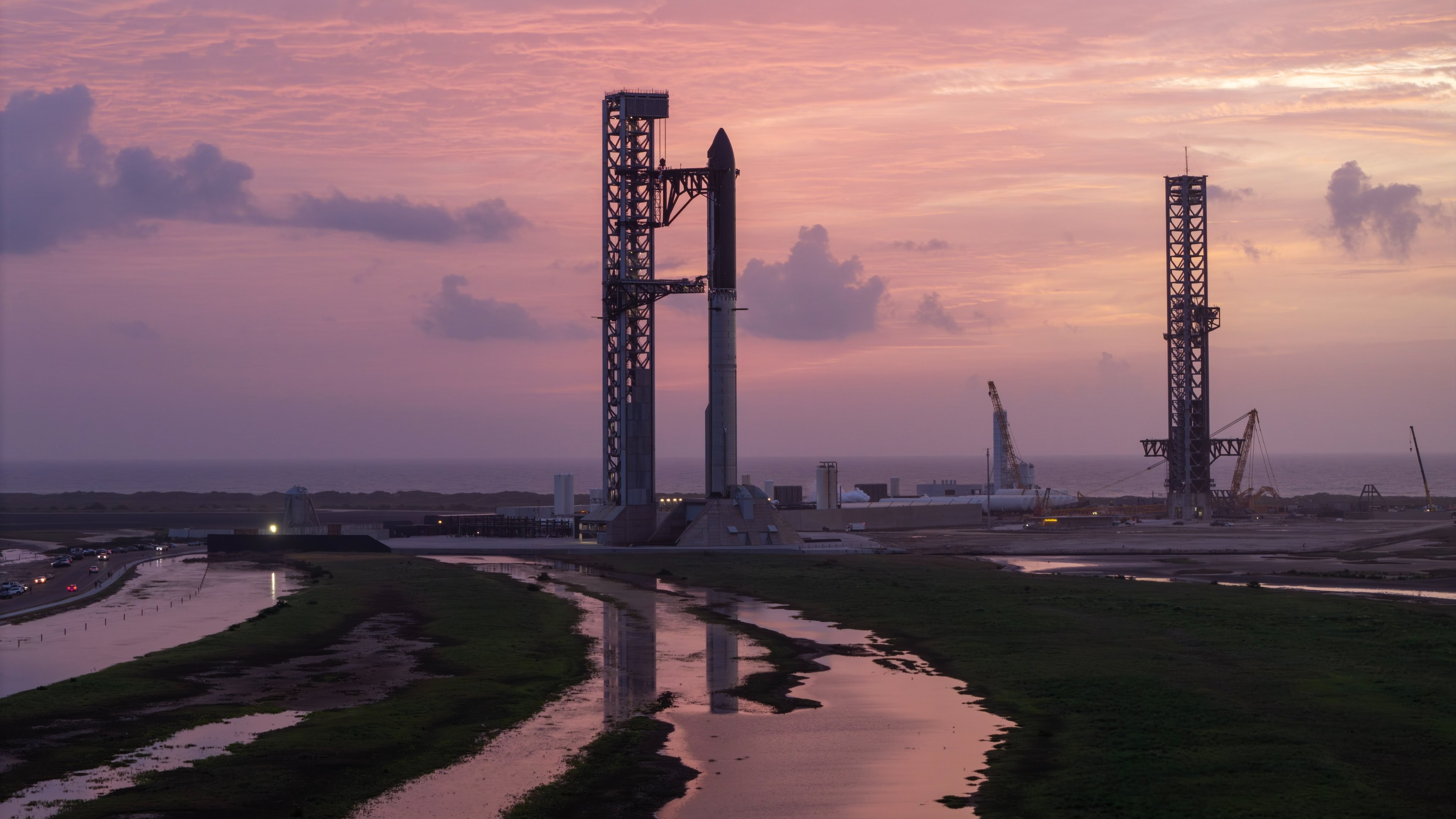 A giant steel rocket stands next to a launch tower reflected in streams of water in the foreground against a rich pink cloudy sky