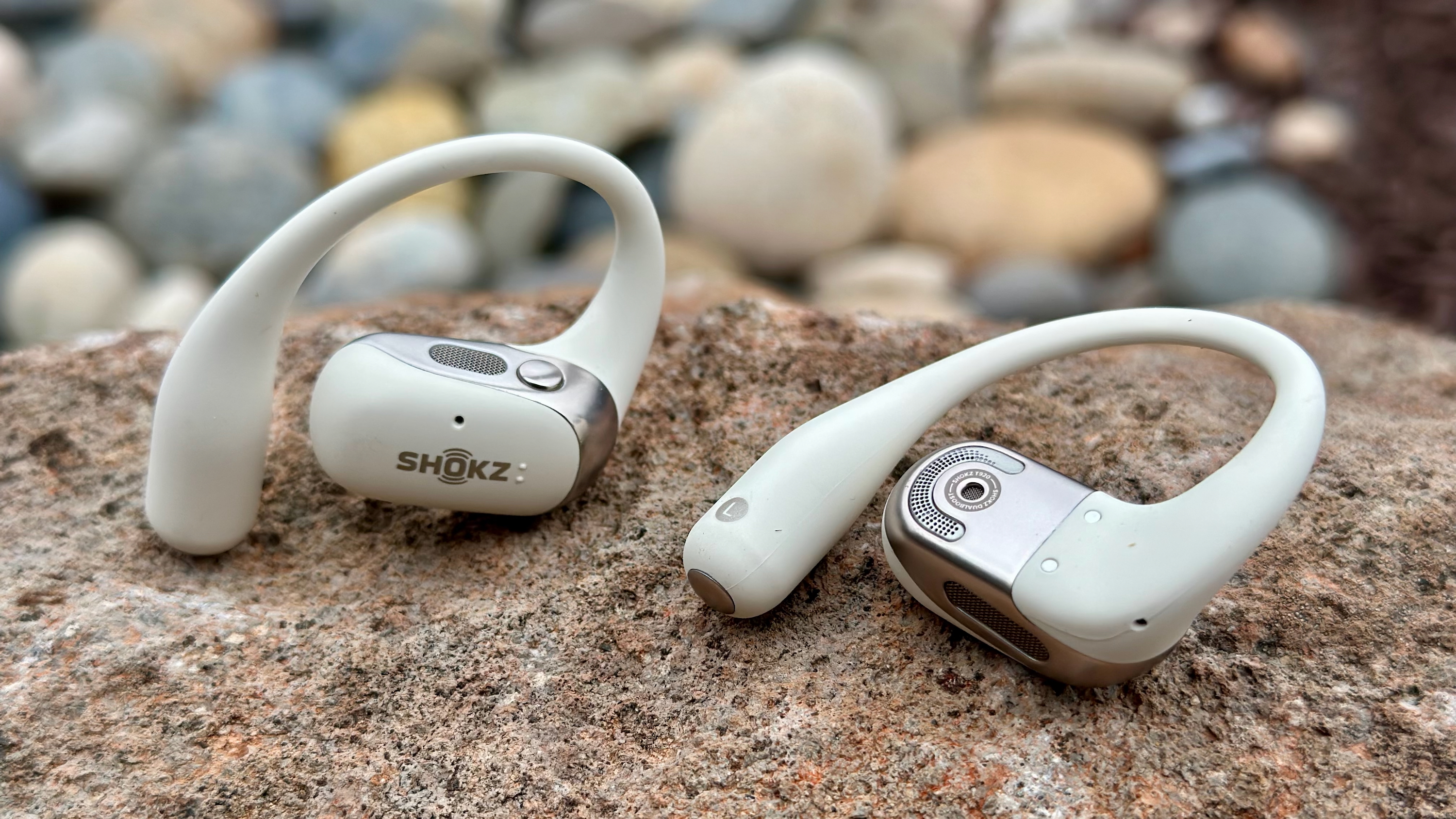 A close-up of the Shokz OpenFit 2 earbuds, one facing the camera showing the logo and top button/speaker grill, the other laying on its back showing the main speaker grill, mic hole, and ear hook.