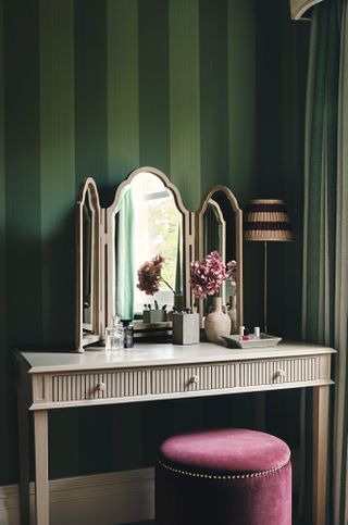 bedroom vanity with matching mirror, plum velvet round stool, two tone green stripe wallpaper, green stripe drapes, lamp and make up on vanity