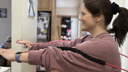 Woman in domestic setting with arms extended in front of her at chest height, holding the ends of a red resistance band