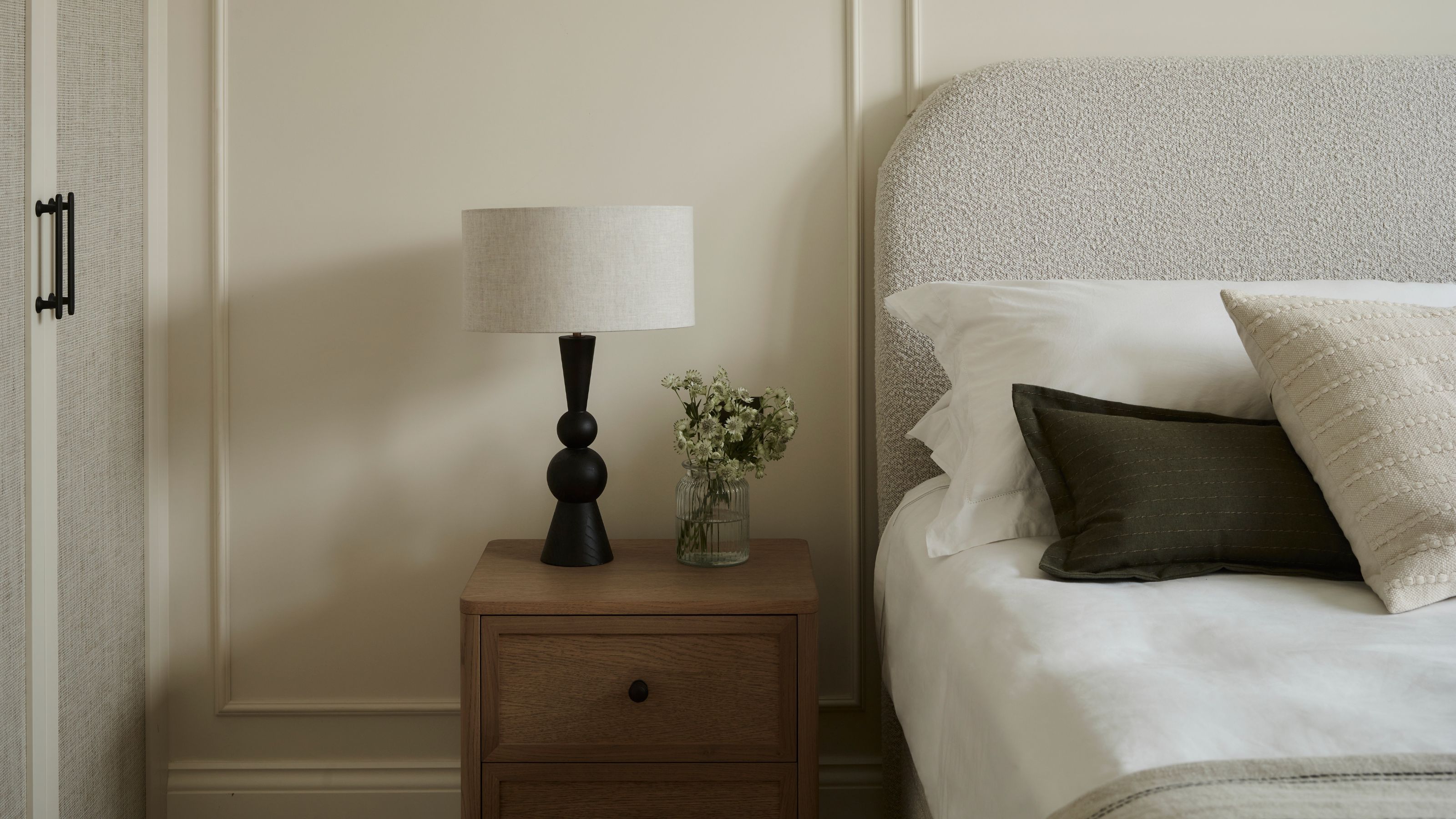 bedroom with timber side table, black table lamp, and vase of flowers, cream walls, a grey headboard, white sheets, olive cushions, and fabric wardrobe doors