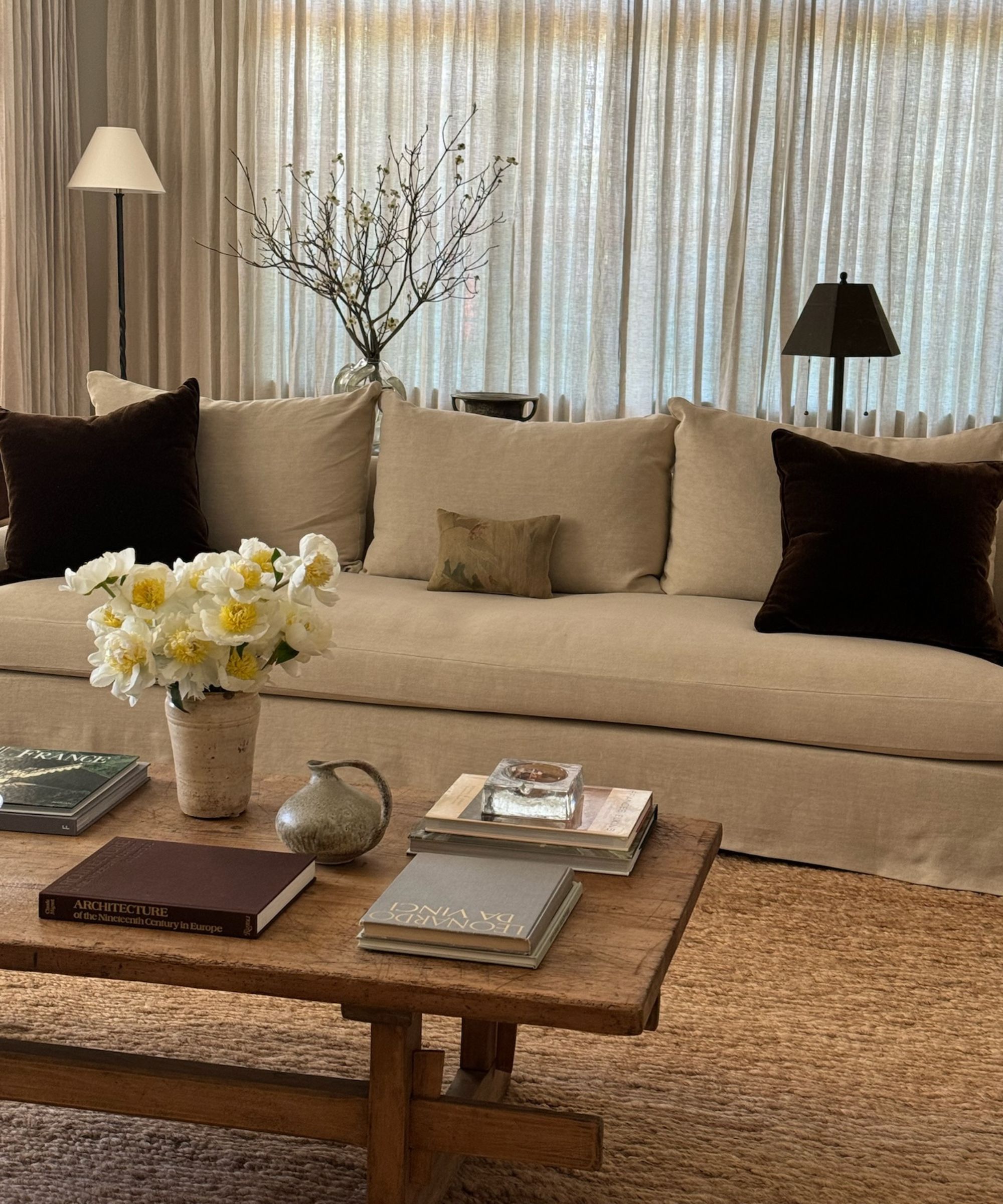 Close up or large beige sofa in a cozy living room with drapes. Textured neutral rug underneath a wooden rectangular coffee table, decorated with books and flowers.