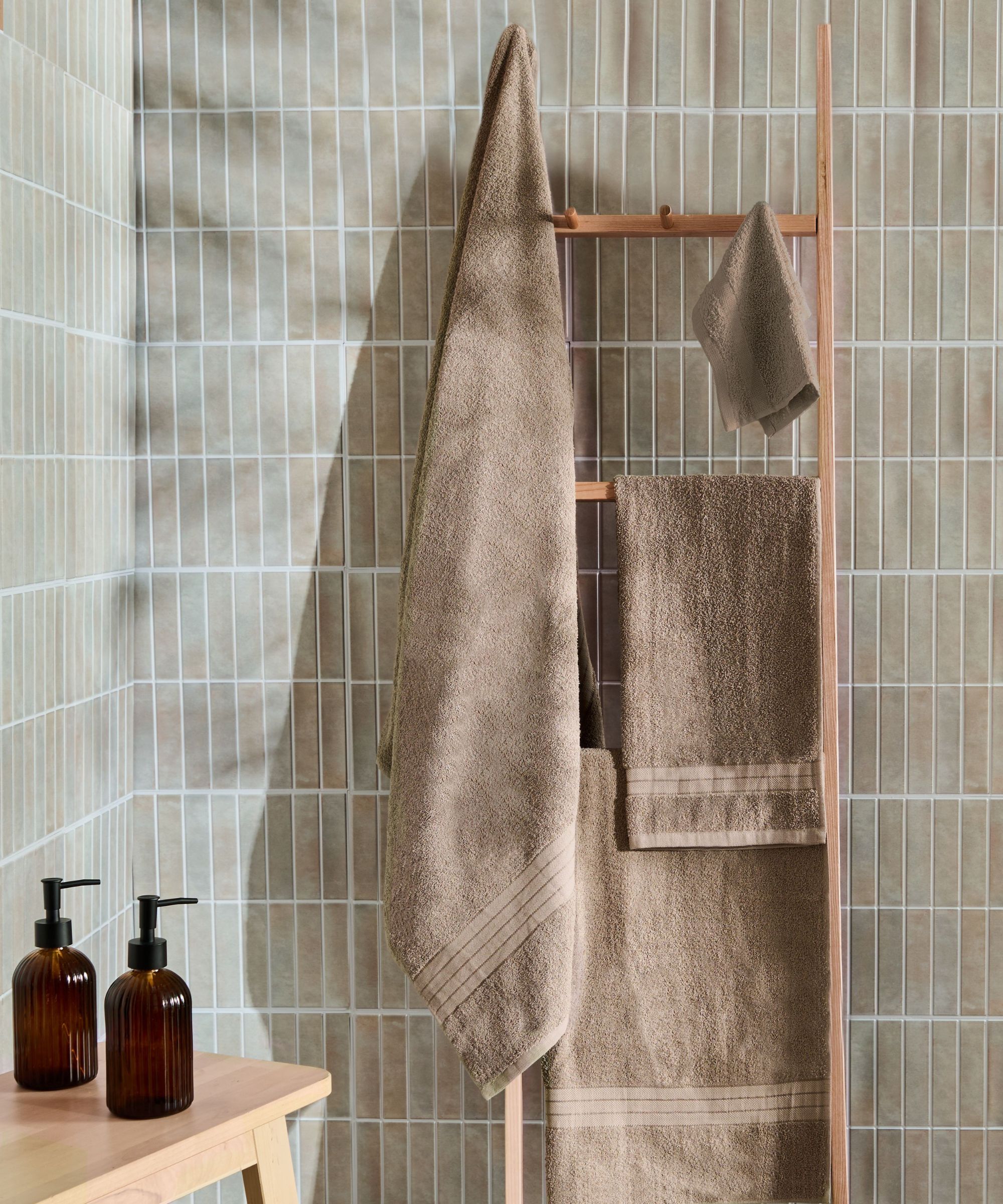 A simple wooden towel ladder leans against soft sage tiles, holding coordinating bath and hand towels in warm taupe. Amber glass pump bottles on a wooden stool add a practical yet stylish finishing touch.