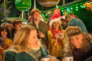 A still from the Beyond Paradise Christmas special showing Humphrey (Kris Marshall) and Martha (Sally Bretton) standing in a crowd at the Shipton Abbott Christmas market. To the left of Humphrey in the crowd are Esther (Zahra Ahmadi) and her daughter Zoe (Melina Sinadinou). There is a Christmas tree at the back of the crowd and large decorations hanging overhead