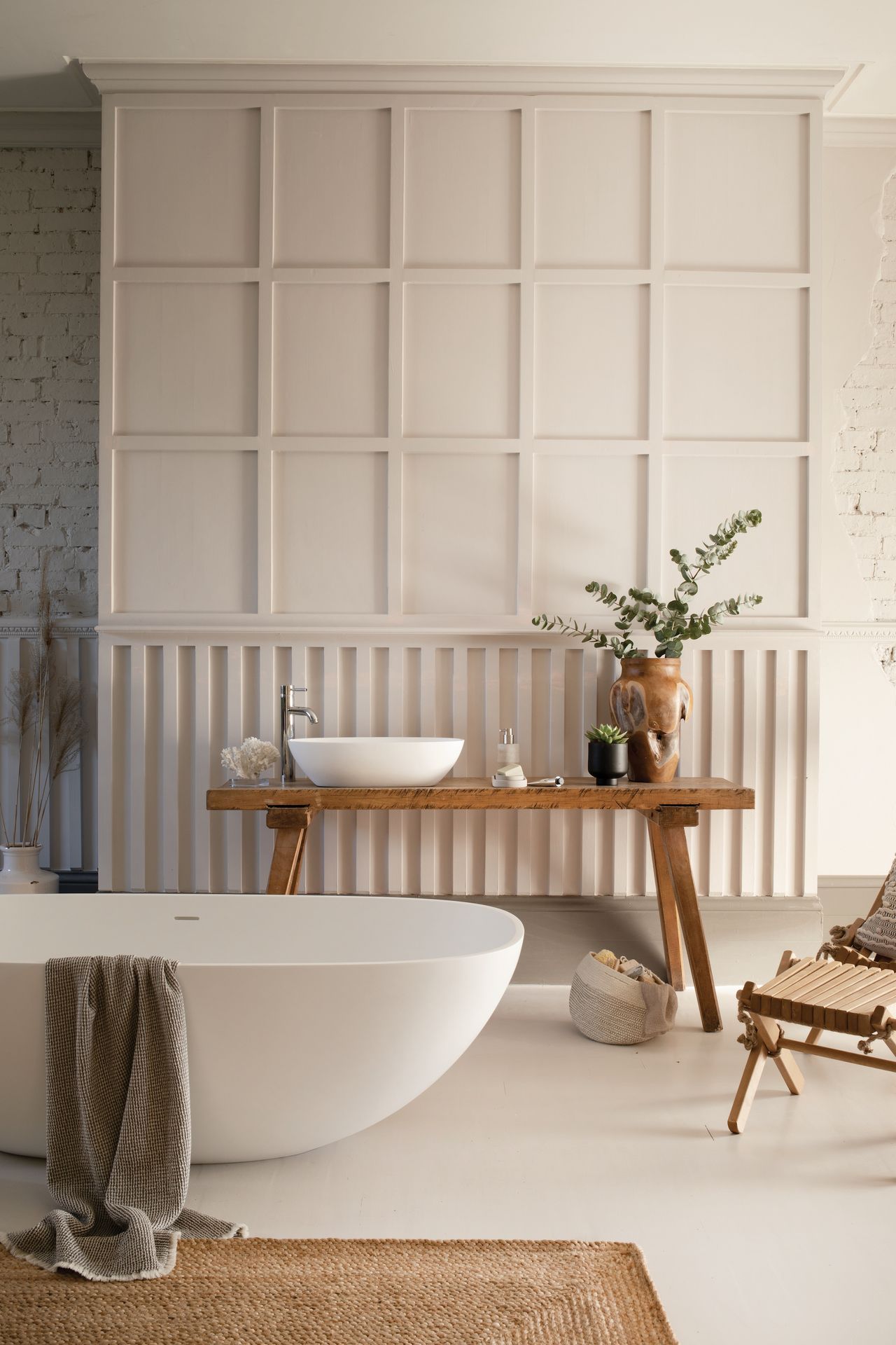 neutral bathroom with modern tub, rustic bench, chair, coir rug, vase, panelled wall, exposed brick wall 