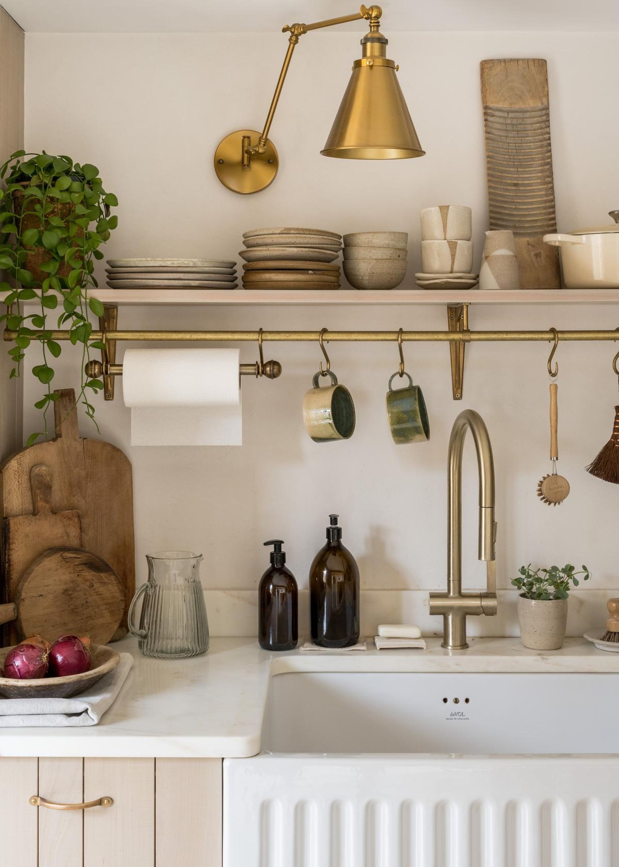 A kitchen with a belfast sink and above the sink is a rack with mugs hanging from it as well as an open shelving unit with ceramic on top of it and a gold brass wall light