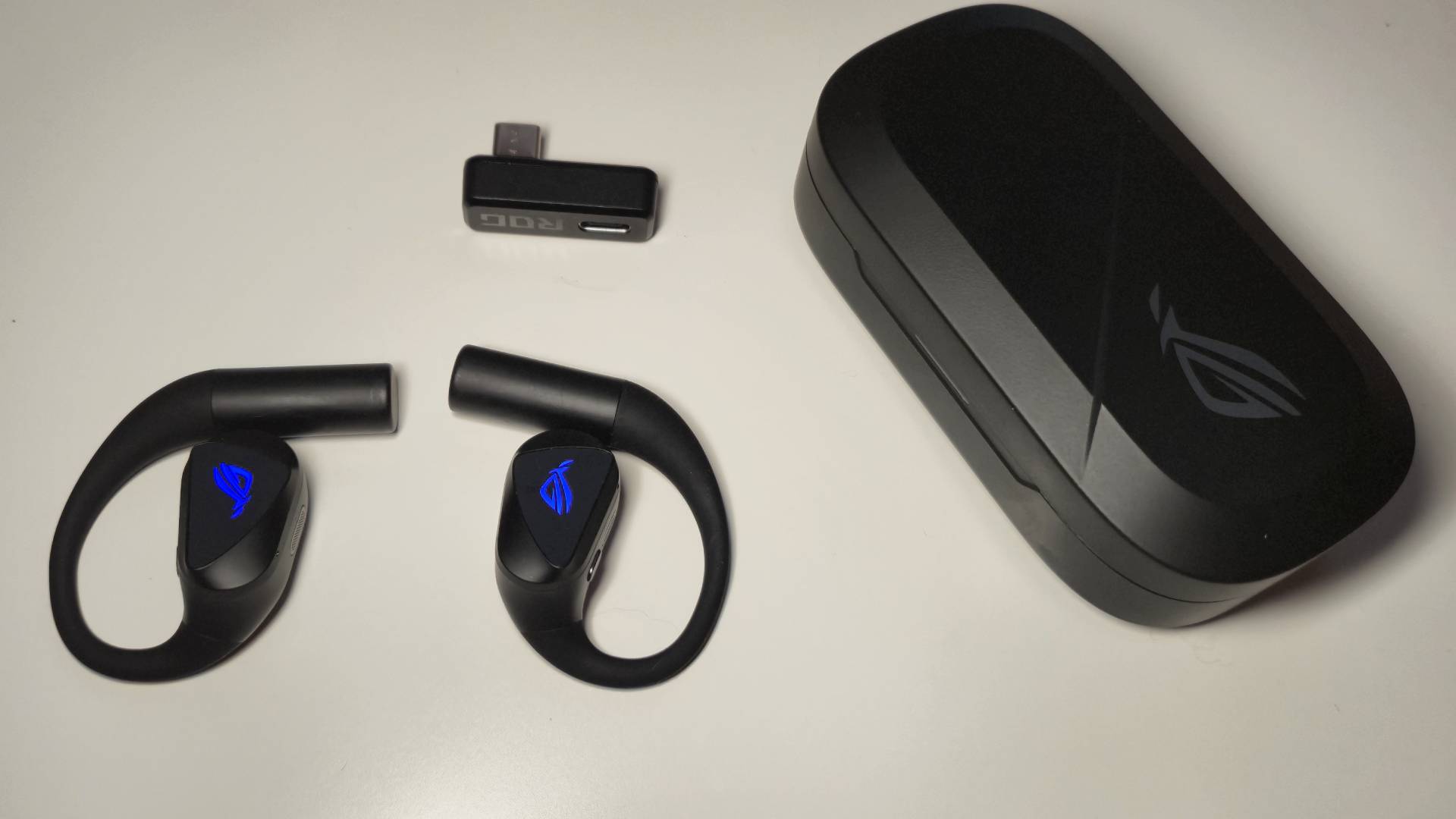 Photo of the Asus ROG Cetra Open Wireless gaming earbuds on a white table with a dongle and case to its side.