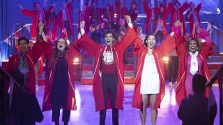 From left to right: Carlos, Ashlyn, Ricky, Gina and Kourtney wearing red graduation gowns and raising their arms up while holding hands.