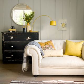 a neutral living room with white panel walls, white couch with yellow cushions and blue striped throw and a distressed black wood chest of drawers
