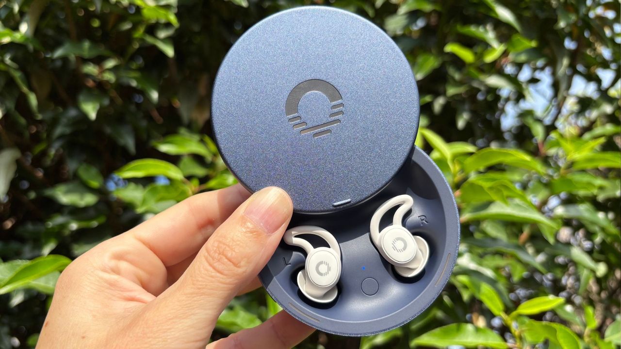 The image shows a hand holding a pair of white Ozlo Sleepbuds 2 in their blue circular case, set against a backdrop of green foliage