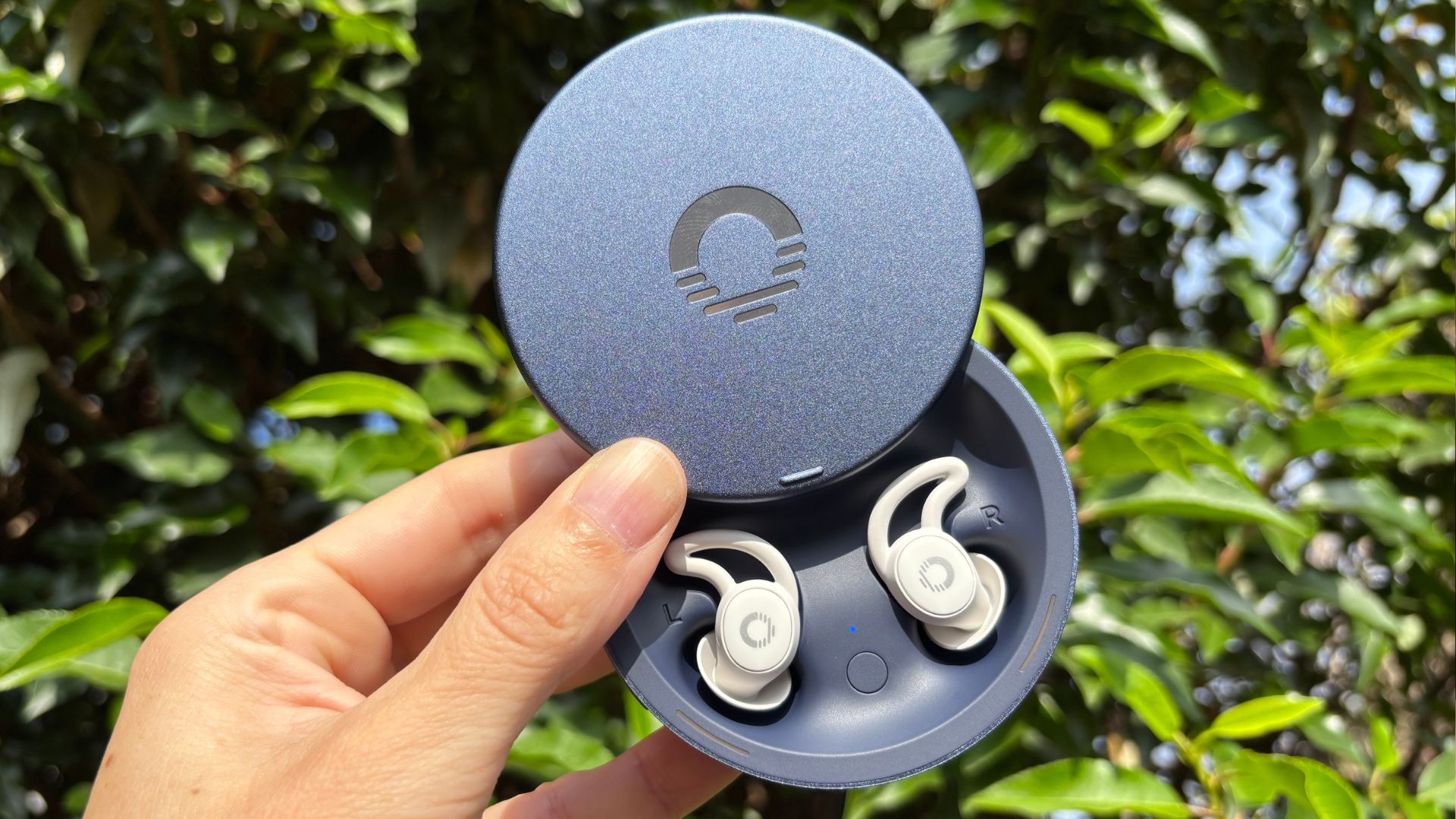 The image shows a hand holding a pair of white Ozlo Sleepbuds 2 in their blue circular case, set against a backdrop of green foliage 