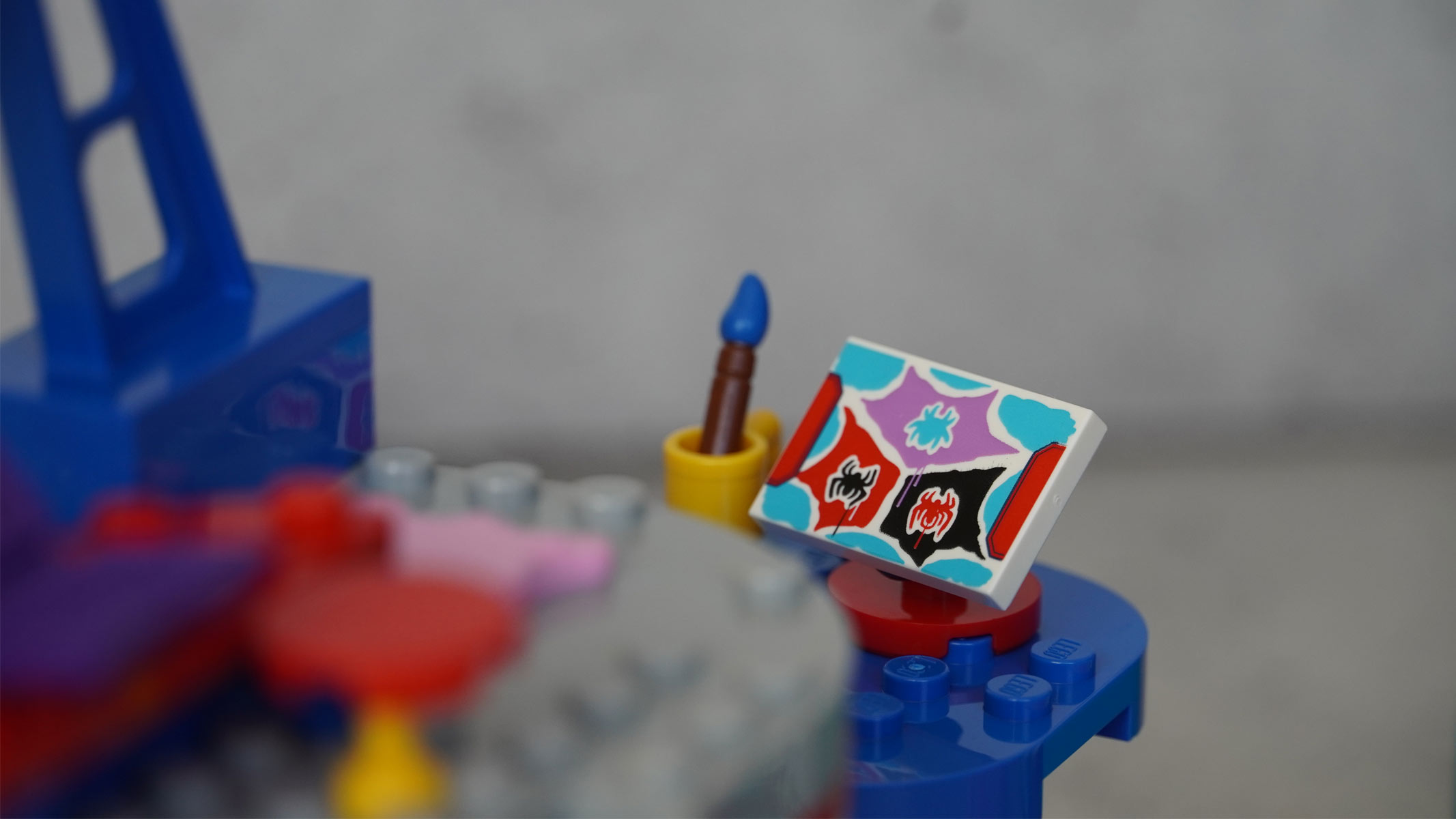 A small square with a multi-colored design is placed next to a tiny paint brush, all in brick form on a grey background.