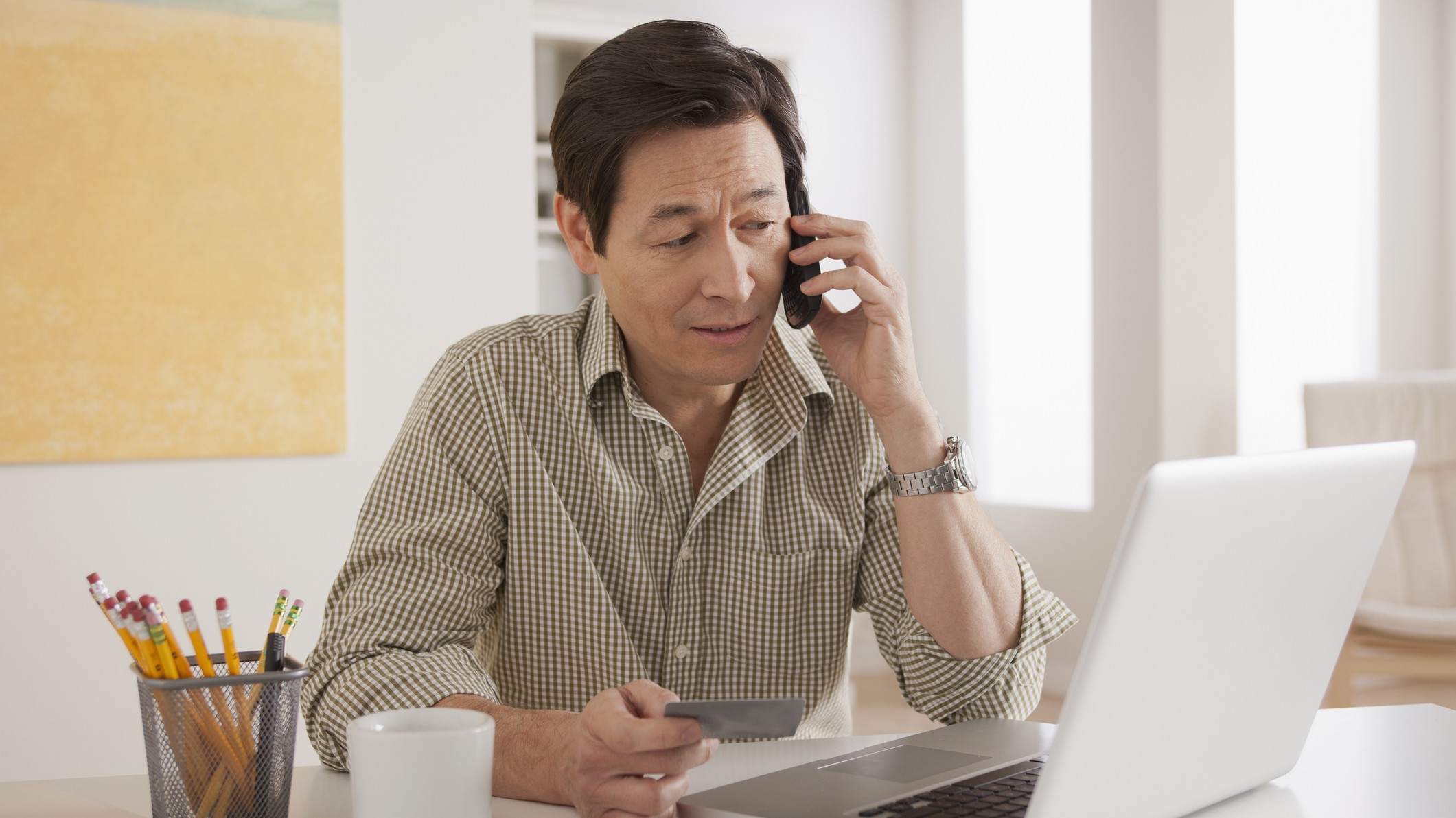 A man makes a phone call while holding a credit card and looking at his computer.