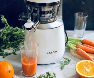 Hurom slow juicer in white on a countertop with carrots and oranges around it