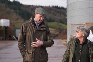 Prince William wearing a green jacket and flap cap looking down and talking to a woman in a green coat who is smiling up at him as they walk in front of green hills and a silo