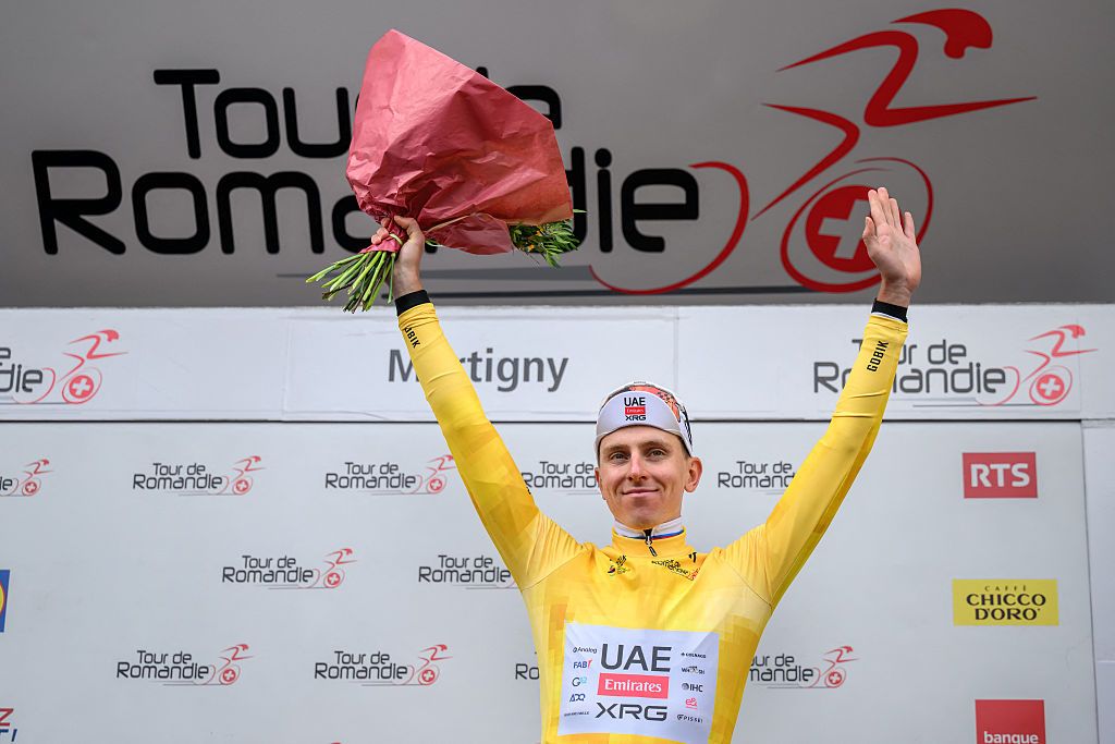 Tadej Pogačar stands on the podium at the Tour de Romandie, wearing the yellow jersey as race leader, with his arms raised in celebration holding a bouquet