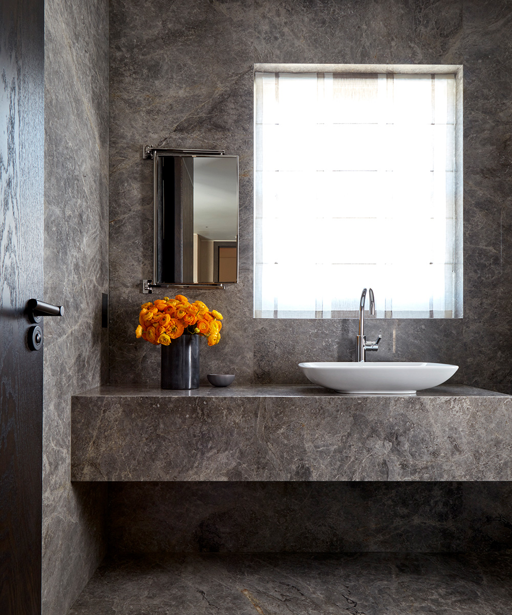 A dark grey stone covered bathroom trend with white sink and a vase of orange flowers.