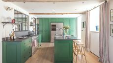 Kitchen and dining area with green cabinets, beamed ceilings, paneled window above gas range, kitchen island with faucet and flower vase next to high stools. On hardwood floors in well-lit room.
