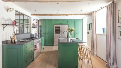 Kitchen and dining area with green cabinets, beamed ceilings, paneled window above gas range, kitchen island with faucet and flower vase next to high stools. On hardwood floors in well-lit room.