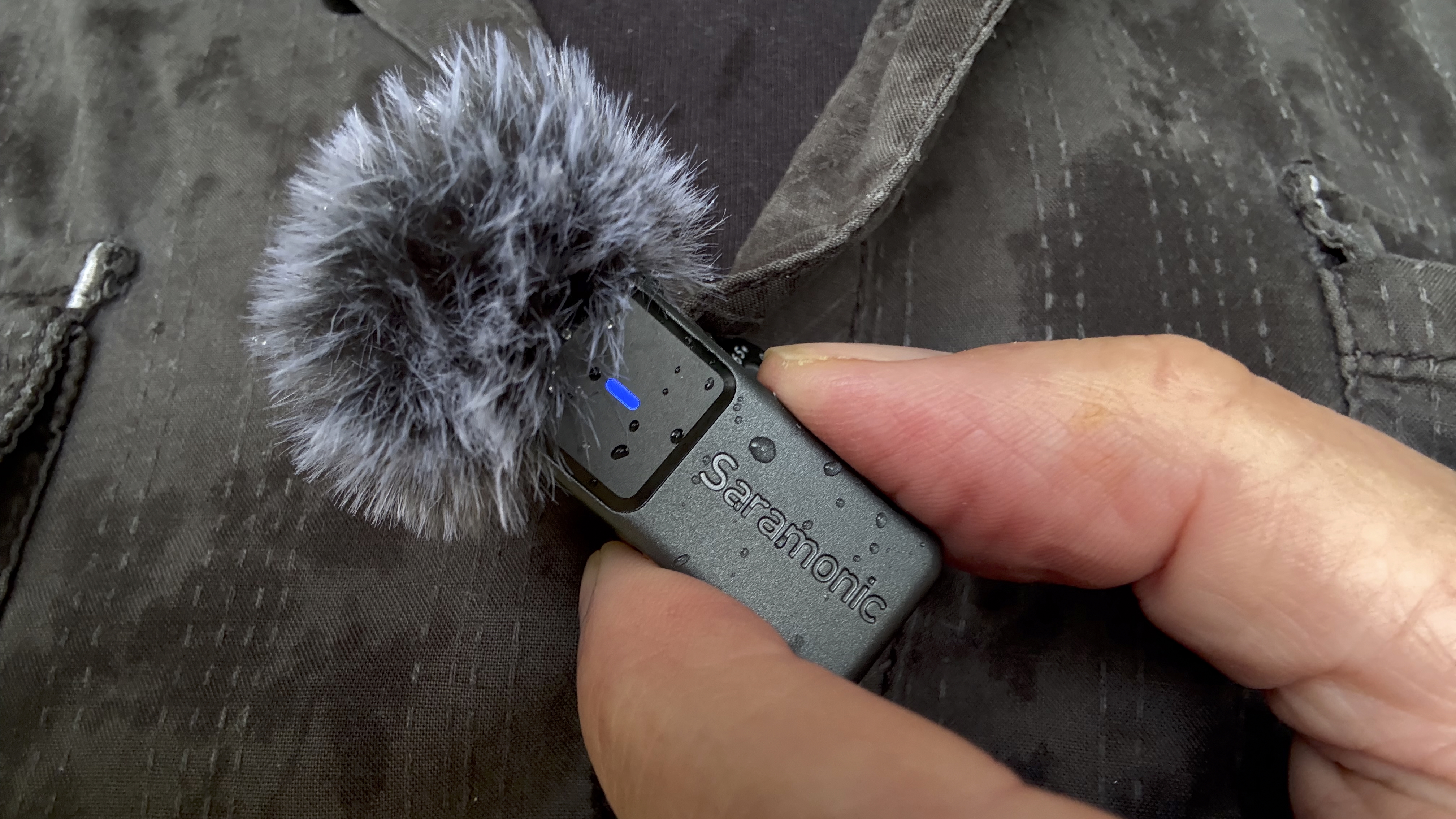 Close-up of a hand holding the transmitter mic which is clipped to a shirt. The mic is ‘wearing’ a fluffy windshield