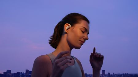 A woman wearing an athletic tanktop and the white Shokz OpenFit 2 wireless earbuds with her eyes closed, listening to the music, with a purple sky and a distant building skyline behind her.