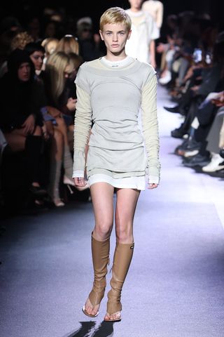 A female model with short hair wearing a long sleeved shirt, a short skirt and knee high leather sandals.