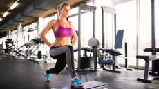 A woman in a gym performs a reverse lunge using a step platform. She wears leggings, a sports bra and sneakers. Her knees are bent, with her right foot resting on the platform in front of her and her left foot behind Her hands rest on her hips as she looks forward; behind her we see various gym machinery.