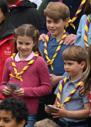 Princess Charlotte wearing a pink sweater standing next to Prince Louis in a blue polo shirt and Prince George in a blue polo shirt, all wearing red-and-yellow scouts scarves