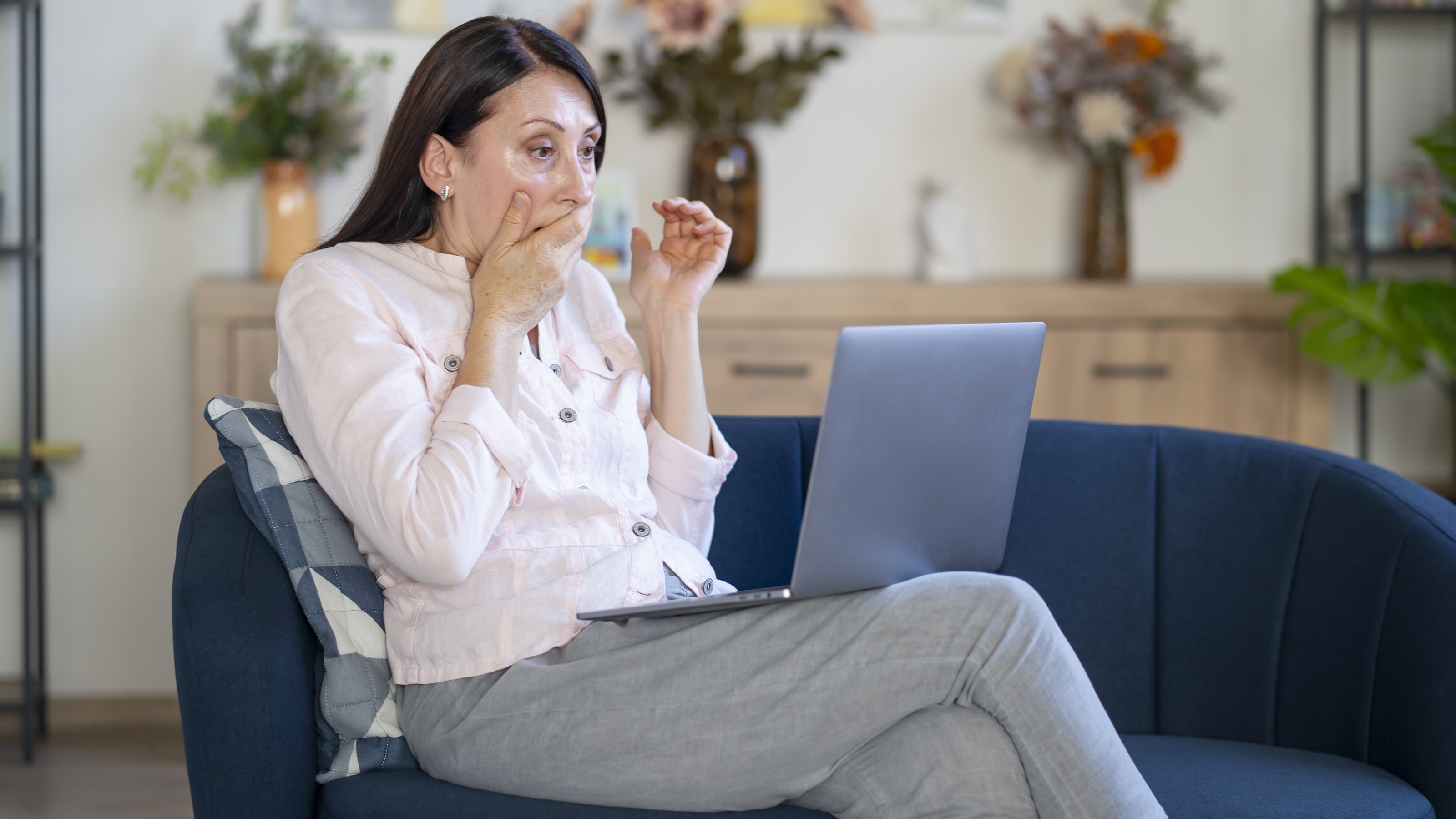 An older woman covers her mouth in shock as she looks at her laptop on her sofa. 